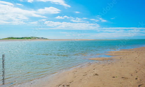 Fototapeta Naklejka Na Ścianę i Meble -  Sunlit waves on the yellow sand of a sunny beach along the North Sea illuminated by the light of a colorful sun and a blue cloudy sky in summer, Walcheren, Zeeland, the Netherlands, July, 2021
