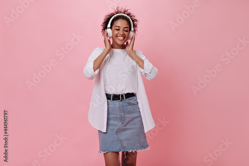 Woman in great mood is listening to music with headphones on pink background. Smiling girl in denim skirt and white shirt posing on isolated backdrop