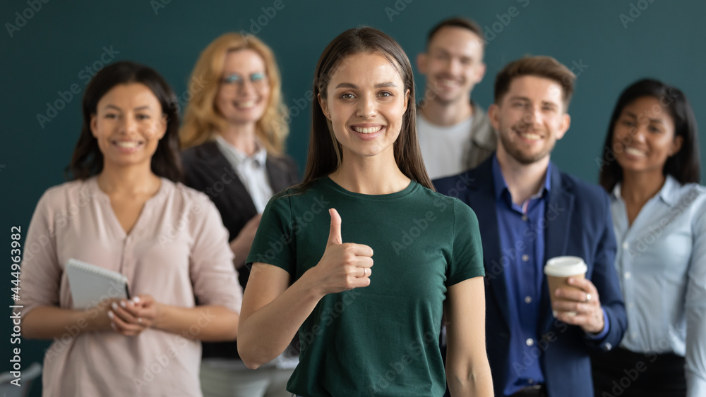 Happy young female business leader making like thumb up, diverse team smiling in background. Satisfied customer, employee with multiethnic group behind giving positive feedback. Head shot portrait