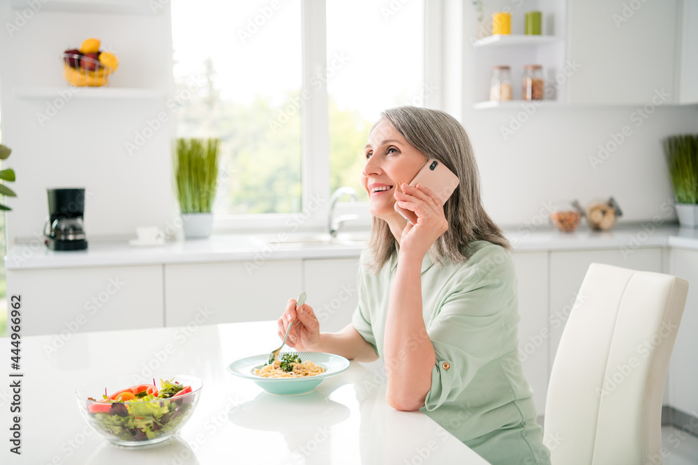 Profile side view portrait of attractive cheerful grey-haired woman eating fresh homemade dinner talking on phone at home light white indoors