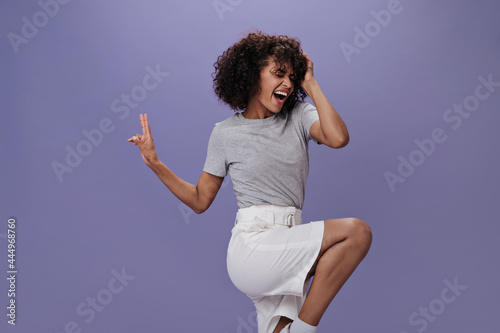 Girl in white shorts jumping, singing and showing peace sign. Optimistic woman in gray t-shirt smailing and dancing on purple backdrop