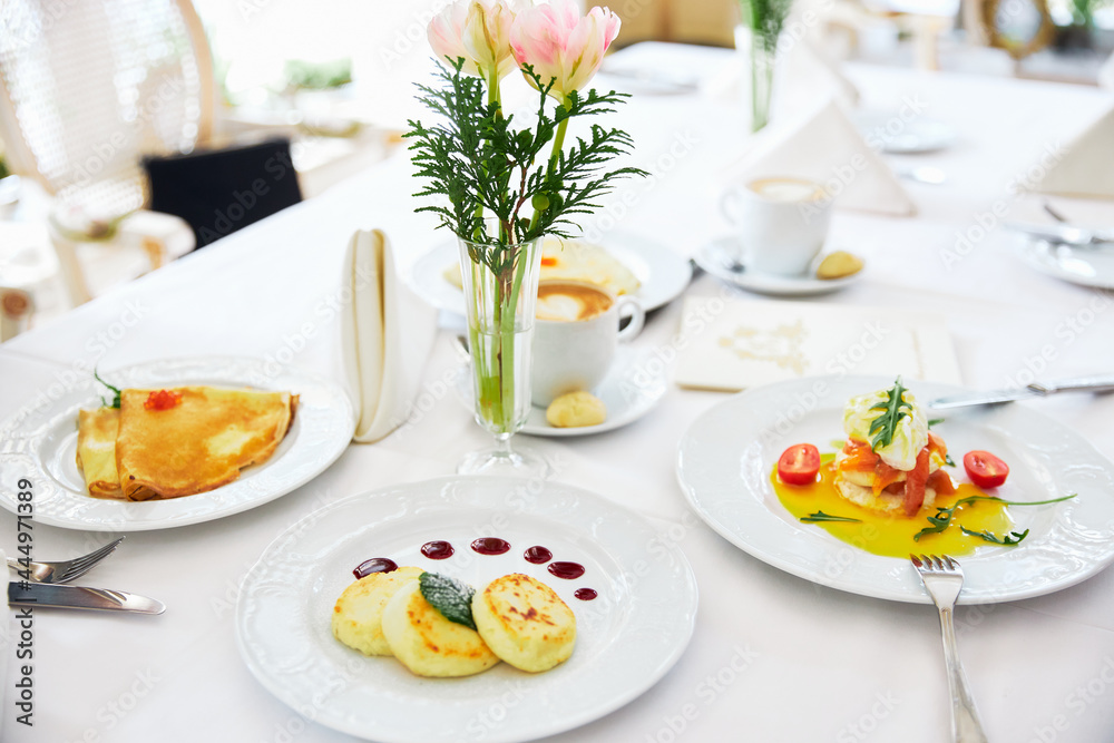 Three cheesecakes and pancakes on a white plate with jam, background of a vase with flowers, restaurant Three cheesecakes and pancakes on a white plate with jam, background of a vase with flowers, restaurant