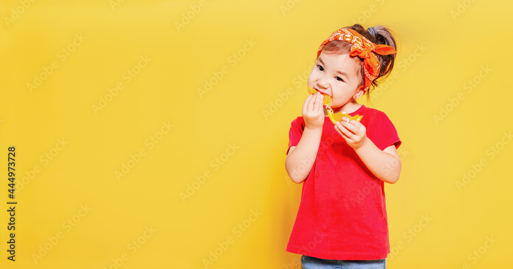 a beautiful little girl in a red T-shirt and an orange bandana eats a slice of orange on a yellow background