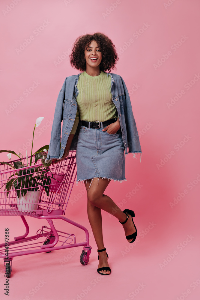 Girl in stylish outfit lifts her leg on pink background and poses with trolley from store. Full-lenght portrait of cheerful woman in denim jacket and skirt