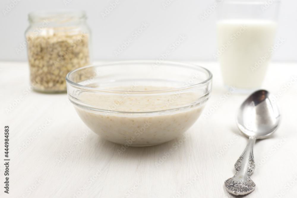 Oatmeal porridge cooked with milk, a glass of milk, a spoon, a plate on a white wooden table Oatmeal porridge cooked with milk, a glass of milk, a spoon, a plate on a white wooden table