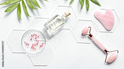 Pink Quarts stone face roller, serum capsules in round glass dish, hexagons, palm leaves. Gua sha stone for beauty facial massage therapy. Natural cosmetics laboratory. Panoramic flat lay, copy-space.