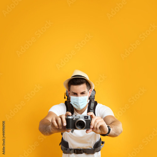 Male tourist in protective mask with travel backpack taking picture against yellow background, focus on camera