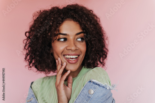 Positive woman in bright top smiling on isolated background. Dark-skinned curly girl in green tee and denim jacket posing on pink backdrop