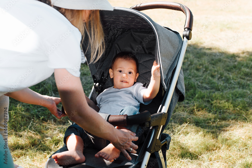 Motherhood and family concept. Young mother seating her baby in a stroller, outdoors. Funny little boy sitting in a stroller and looking at the camera.