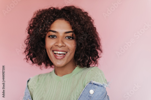 Brown-eyed girl in green T-shirt with smile posing on pink background. Close-up portrait of curly woman in deni, jacket looking into camera on isolated backdrop