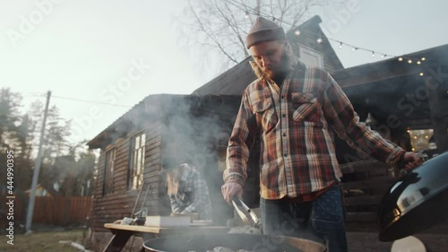 Low angle shot of man taking off lid frim bbq grill and using tongues for turning meat while cooking food outdoors in backyard photo