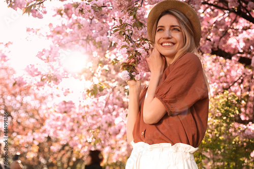 Young woman wearing stylish outfit near blossoming sakura in park. Fashionable spring look