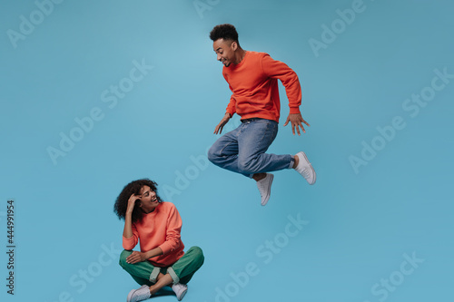 Happy girl in orange sweatshirt sitting. Man jumping on blue background. Portrait of guy in jeans moving and girl chilling on isolated