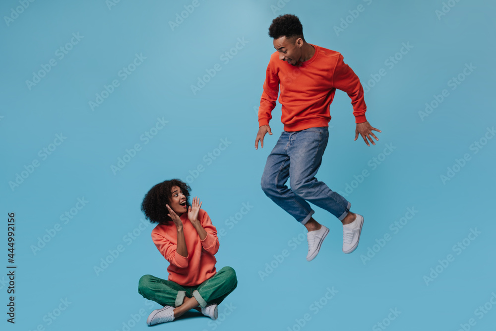Curly woman sit while her boyfriend jumping on blue background. Portrait of brunette man in movement. Guy in jeans and girl in orange sweater pose on isolated