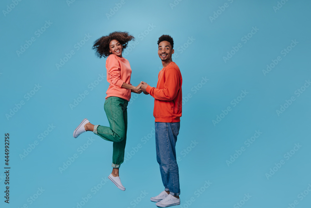 Couple holds hands and smiles on blue background. Curly brunette woman in green pants jumps and dark-skinned man looks into camera Couple holds hands and smiles on blue background. Curly brunette woman in green pants jumps and dark-skinned man looks into camera