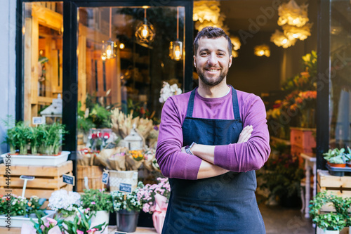 Flower shop owner in an apron has his arms crossed in front of his flower store.