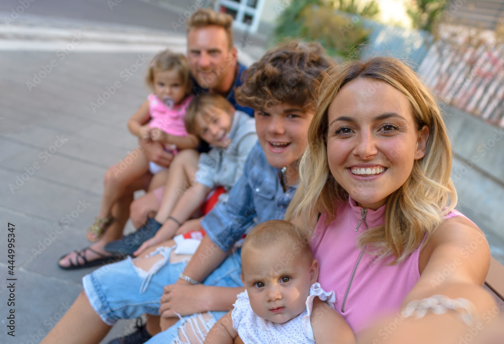 young adult family time having fun outdoors sitting on a bench. mom and dad portrait on holidays with children visiting urban city centre. happy selfie on vacation days. joy and lifestyle concept