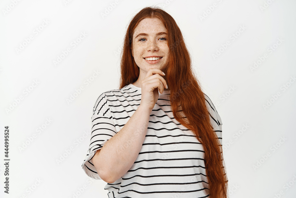 Image of beautiful confident redhead woman with long natural hairstyle, freckles, smiling pleased, touch chin and look with thoughtful face expression at camera, white background Image of beautiful confident redhead woman with long natural hairstyle, freckles, smiling pleased, touch chin and look with thoughtful face expression at camera, white background