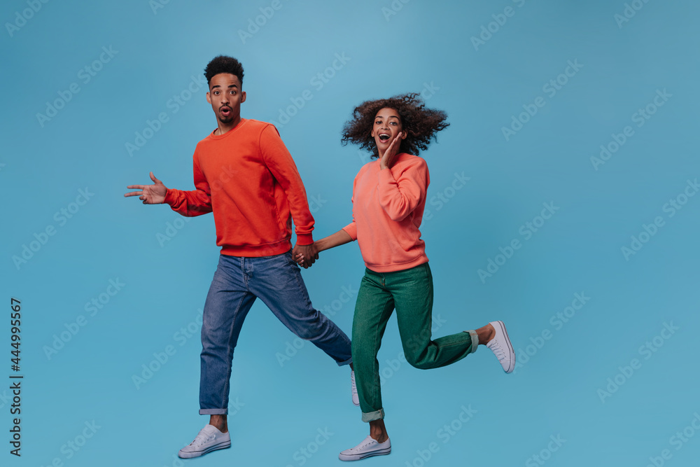 Man and woman look surprised into camera and run on blue background. Brunette girl in green pants and man in red sweater move on isolated Man and woman look surprised into camera and run on blue background. Brunette girl in green pants and man in red sweater move on isolated