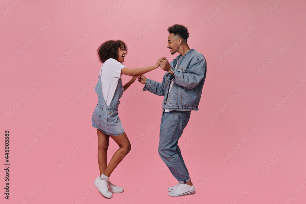 Positive man and woman in denim clothes dancing on pink background. Charming guy and dark-skinned girl in jeans moce on isolated