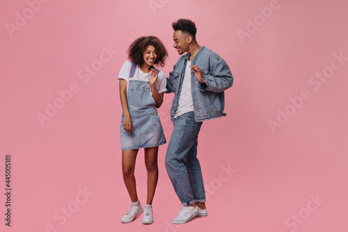 Joyful man and woman in denim outfits smiling and posing on pink background. Dark-skinned guy in jeans and pretty girl dance on isolated