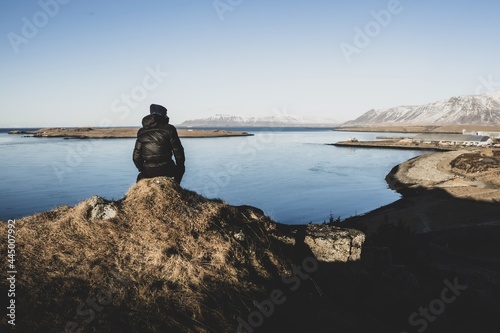 A man in a winter jacket sitting on a rock, watching a winter panorama of snowy Mount Esja ridge and Atlantic ocean. Reykjavik, Iceland.  photo