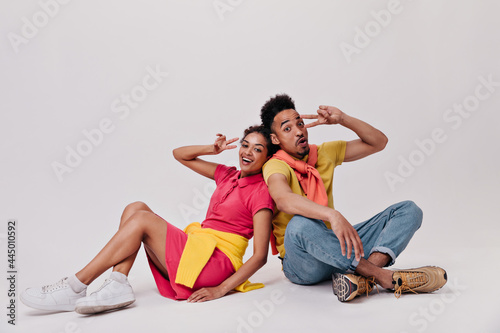 Happy man and woman in colorful clothes showing peace signs. Attractive brunette girl in sport-style dress and guy in jeand and yellow tee smile on isolated photo