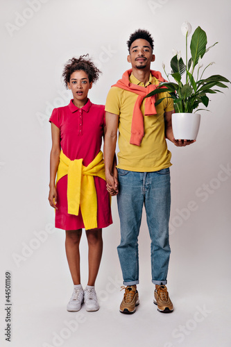 Full length portrait of man and woman in colorful outfits look into camera and hold plant. Brunette guy on yellow tee and pretty woman in red dress pose on isolated