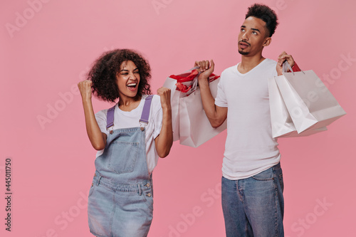 Man in Bad mood holding shopping bags. Woman enjoys shopping. Brunette girl in denim dress smiles and guy in jeans looks dissatistied photo