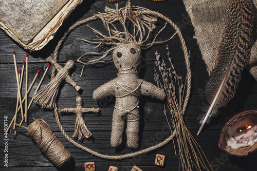 Female voodoo doll with pins surrounded by ceremonial items on black wooden background, flat lay photo