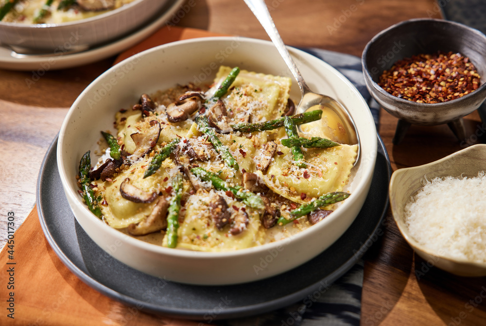 Homemade mushroom ravioli with asparagus in a bowl on a wooden table with a beautiful rust colored linen beneath it. Bowl has red pepper flakes and parmigiana in small bowls next to it. Homemade mushroom ravioli with asparagus in a bowl on a wooden table with a beautiful rust colored linen beneath it. Bowl has red pepper flakes and parmigiana in small bowls next to it.