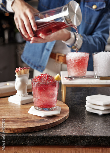 Woman pours a pomegranate granita in background. Frosty granita in foreground on a white coaster on top of a wooden surface.  Jonathan Adler Mohawk match holder behind the drink.