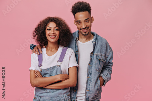 Happy dark skinned man and woman smiling and hugging on pink background. Cool brunette curly girl in white tee and guy in denim outfit laughing on isolated