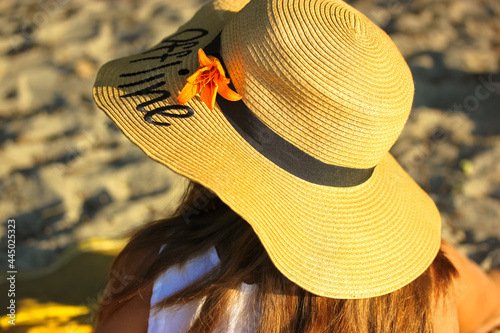 A girl in a straw hat with a word Offline written on it  adorned with an orange lily is resting on the seashore on a sunny summer day. View from the back. Concept of vacation  summer vacation  travel.