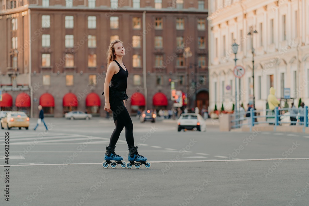 Obraz premium Full length shot of slim dark haired woman rollerblades on asphalt road dresed in black sportsclothes enjoys outdoor activities exercises for healthy and strong body. Sport and hobby concept