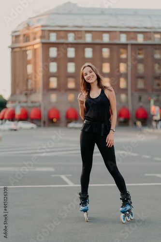 Full length shot of active smiling woman enjoys extreme sport rides on rollerblades dressed in black comfortable clothes stands outdoors learns rollerskating. People leisure recreation concept
