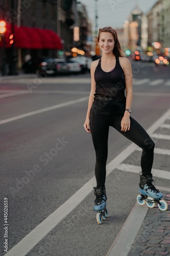 Full length shot of active young woman being in good physical shape dressed in black activewear enjoys rollerblading during good summer day poses on road against city background. Recreation.