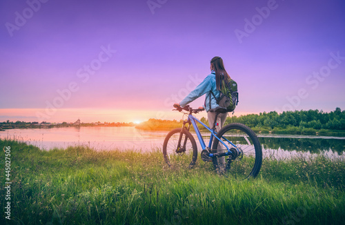 Woman with bicycle near the river at sunset in spring. Landscape with sporty girl with backpack riding a mountain bike, dirt road, green grass, water, purple sky in summer. Sport and travel. Cycle