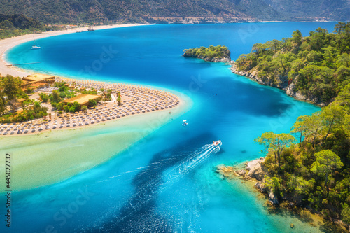 Aerial view of sandy beach, blue sea and speed boat at sunrise in summer. Motorboat in Blue Lagoon, sun beds, clear azure water. Tropical landscape with yacht, green forest. Top view. Oludeniz, Turkey