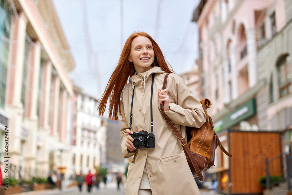 Obraz premium Happy female in beige coat with leather brown backpack taking photo with retro film camera on background of historic building. City traveling alone concept, trip. Copy space