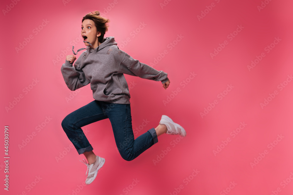 Woman in grey hoodie and jeans jumps on pink background. Emotional teen girl in sweatshirt and denim pants moves on isolated Woman in grey hoodie and jeans jumps on pink background. Emotional teen girl in sweatshirt and denim pants moves on isolated