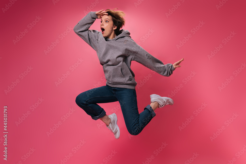 Surprised woman in grey hoodie jumping on pink background. Shocked short-haired girl in jeans moves and dances on isolated backdrop