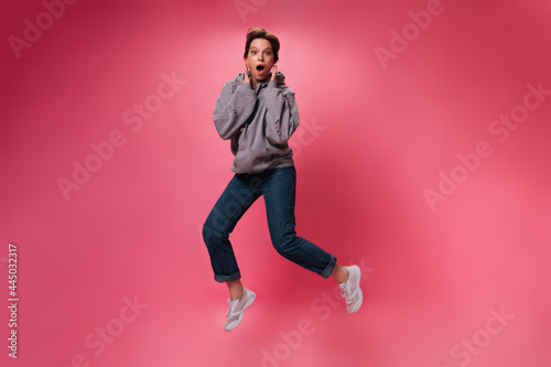 Surprised woman in grey hoodie jumping on pink background. Shocked girl in denim pants and sweatshirt moves on isolated backdrop