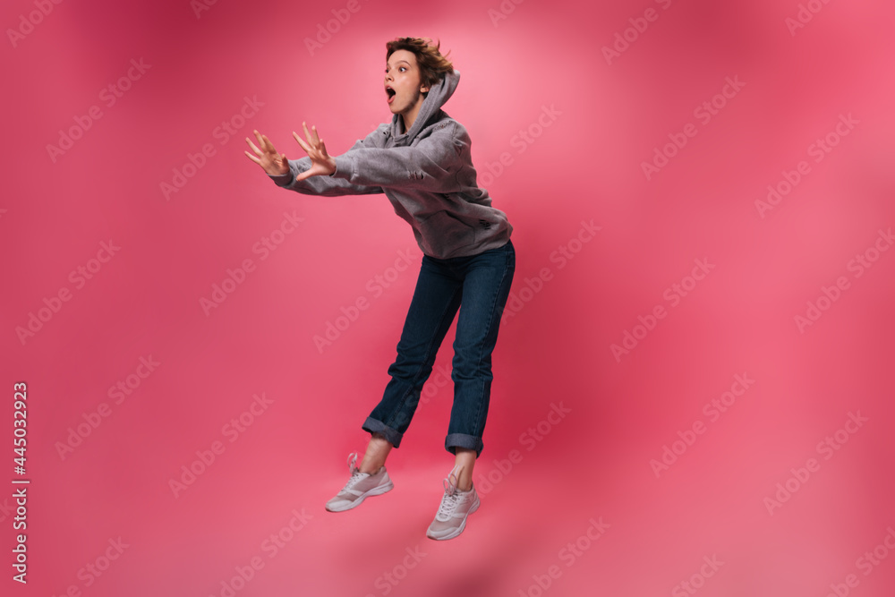 Woman in hoodie and jeans looks surprised and jumps on isolated background. Emotional shocked girl in denim pants moves on pink backdrop