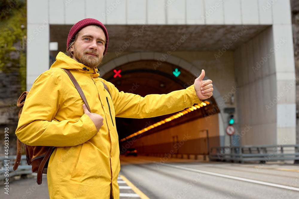 Portrait of young man hitchhiking around the country, trying to catch passing car for traveling. Caucasian hipster man in yellow coat with backpack went hitchhiking to south.