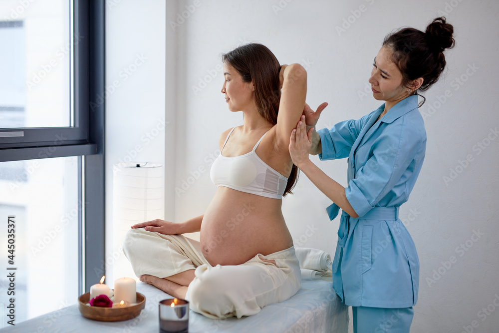 Fototapeta premium Pleasant Female Physiotherapist Massaging Back And Shoulders Of Pregnant Woman Sitting On Bed In Bright Spa Cabinet, With Candles. Adult Mom-to-be Is Enjoying Getting Massage