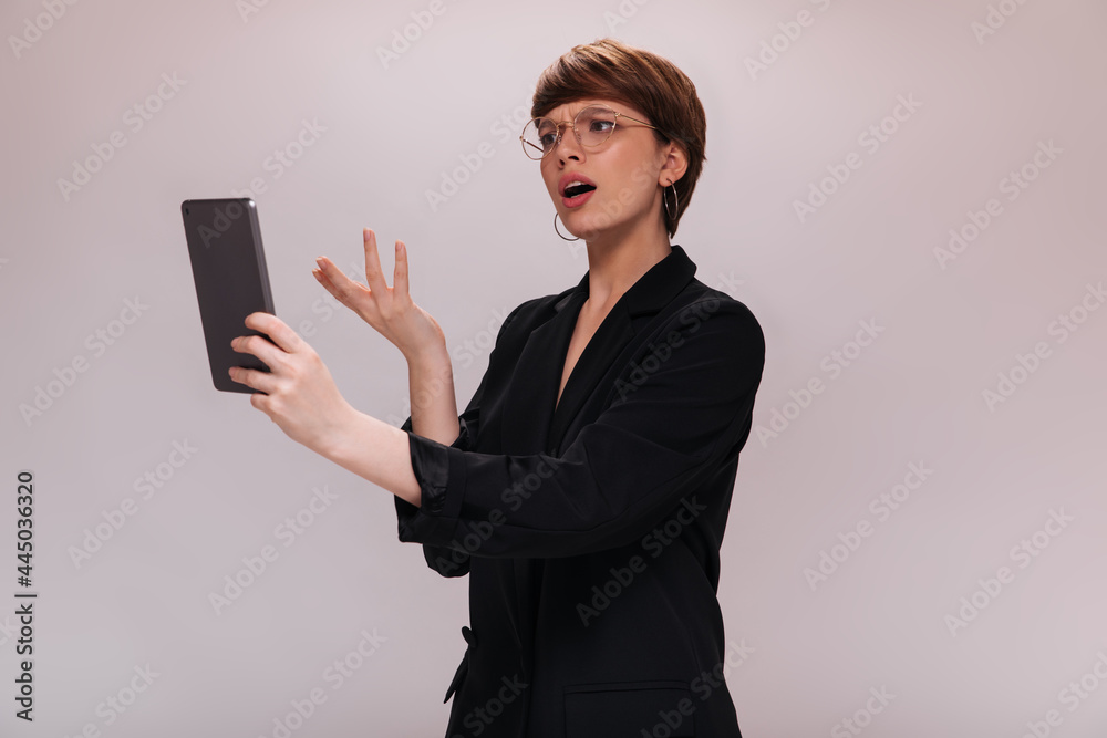 Woman looks into tablet screen with misunderstanding. Business lady in black jacket poses on isolated white background Woman looks into tablet screen with misunderstanding. Business lady in black jacket poses on isolated white background