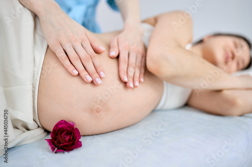 Cropped masseur doing massage to pregnant woman lying on bed relaxed, focus on hands of therapist. Chinese lady in bra get pleasure by massage. pregnancy concept. focus on hands massaging tummy