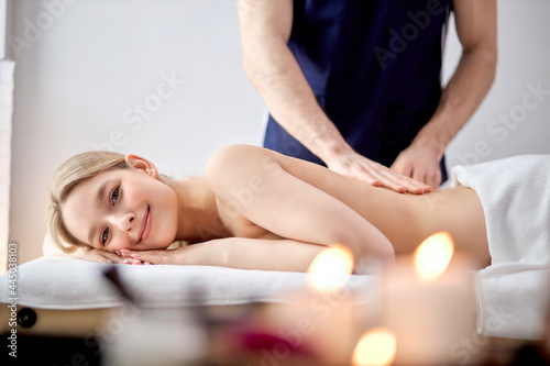 Male hands of massage therapist makes light massage to pretty female in cosmetology room, relaxed shirtless blonde caucasian woman lying on belly getting pleasure, in room with candles