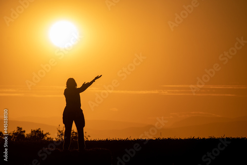 silhouette of a woman in a field with her arms outstretched as if offering something with a large sun and mountains in the background